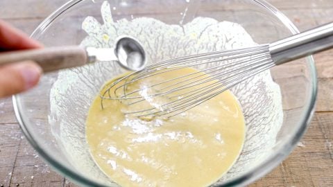 A hand adds baking soda to a bowl of yellow batter with a whisk on a wooden surface.