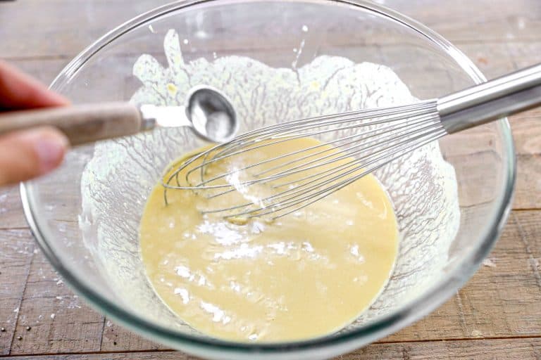 A hand adds baking soda to a bowl of yellow batter with a whisk on a wooden surface.