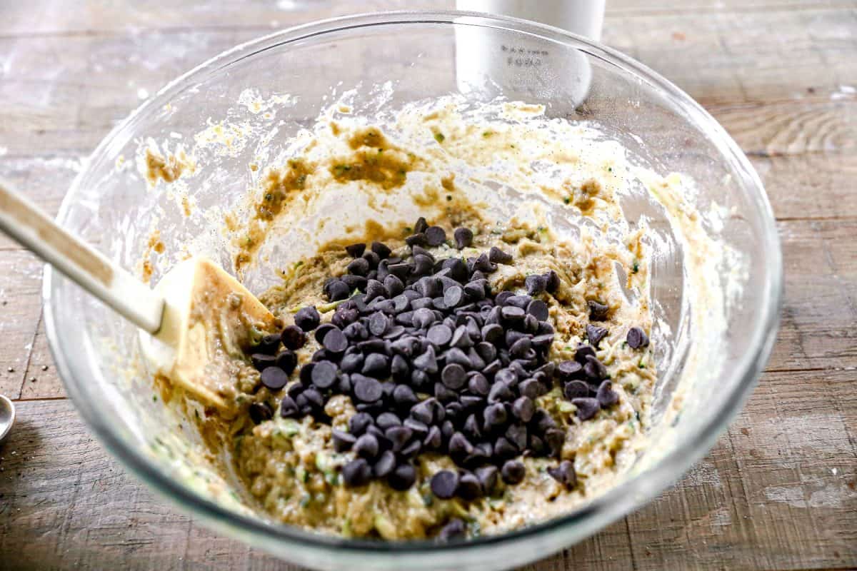 A glass bowl of zucchini bread batter with chocolate chips and a spatula on a wooden surface.