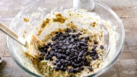 A glass bowl of zucchini bread batter with chocolate chips and a spatula on a wooden surface.