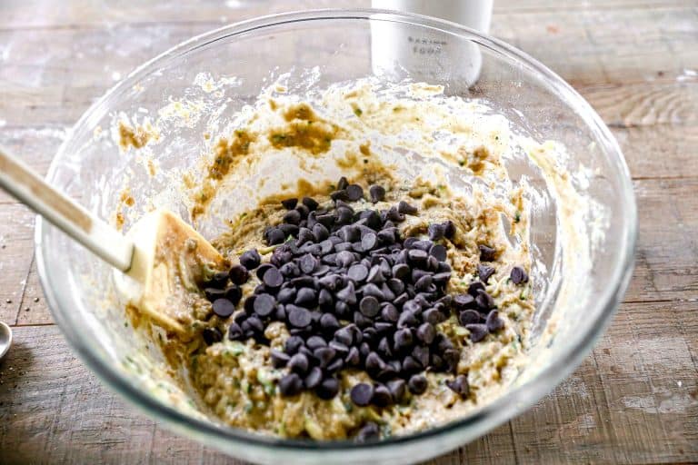 A glass bowl of zucchini bread batter with chocolate chips and a spatula on a wooden surface.