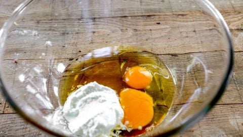 Two raw eggs and a dollop of white yogurt in a clear glass bowl on a wooden surface.