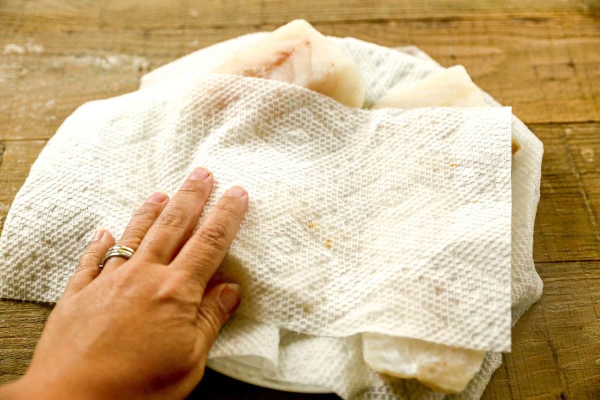 A hand using a paper towel to pat dry raw cod fillets on a wooden surface.