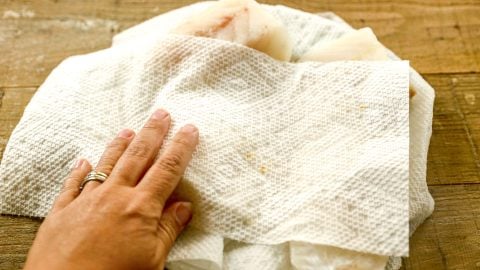 A hand using a paper towel to pat dry raw cod fillets on a wooden surface.