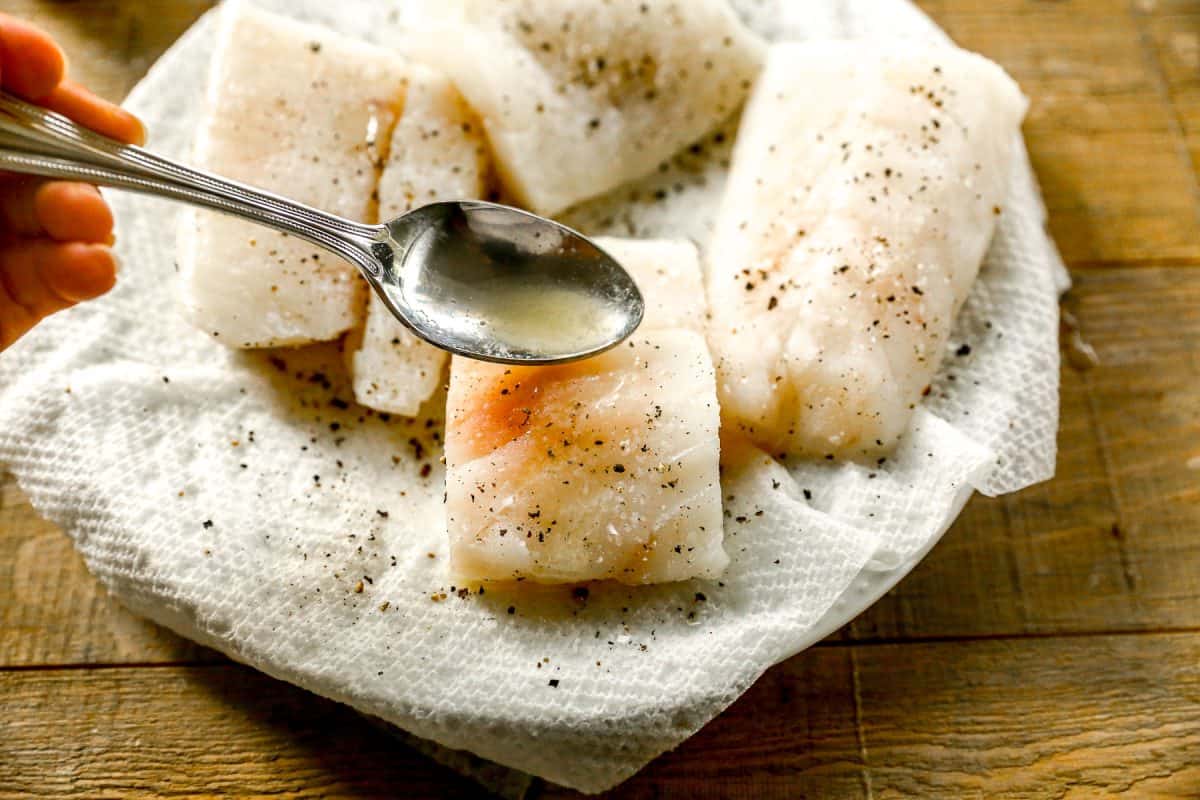 A spoon drizzling liquid over cod fillets seasoned with salt and pepper on a paper towel.