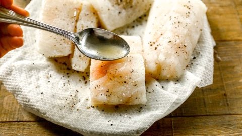 A spoon drizzling liquid over cod fillets seasoned with salt and pepper on a paper towel.
