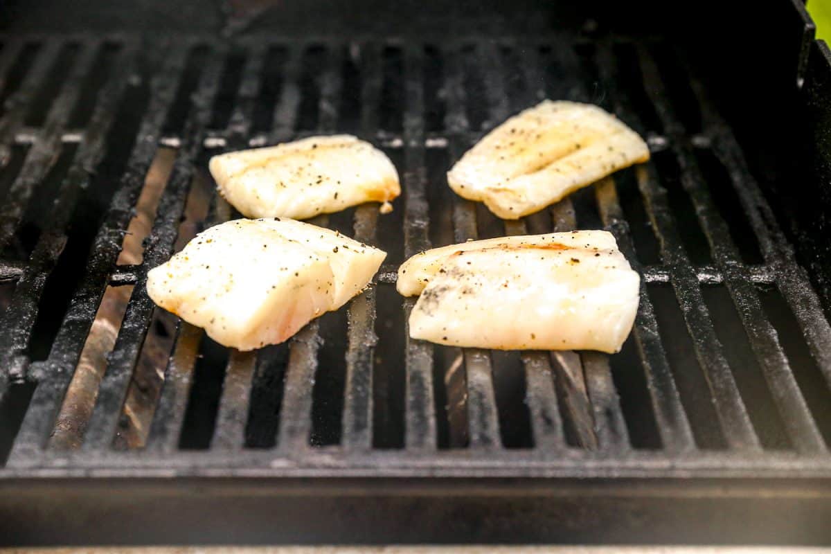 Four seasoned cod fillets grilling on a barbecue with black grates.