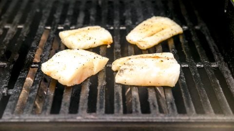 Four seasoned cod fillets grilling on a barbecue with black grates.