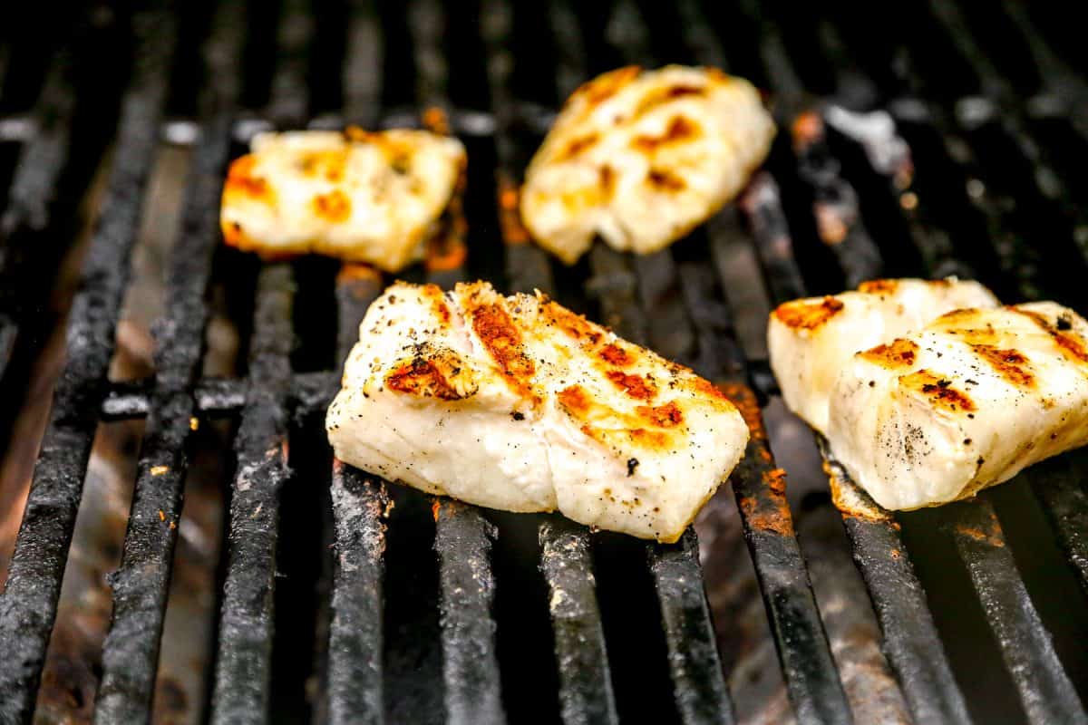 Several pieces of seasoned cod are grilling on a barbecue with blackened grill grates.