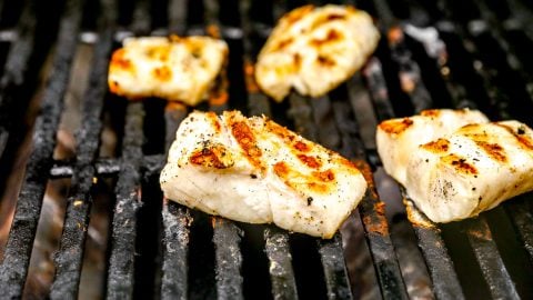 Several pieces of seasoned cod are grilling on a barbecue with blackened grill grates.