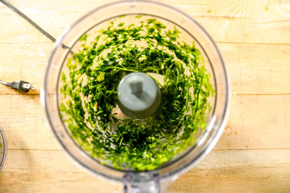 Chopped green herbs in a food processor bowl on a wooden countertop, viewed from above.