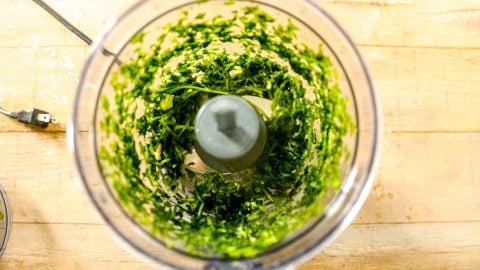 Chopped green herbs in a food processor bowl on a wooden countertop, viewed from above.