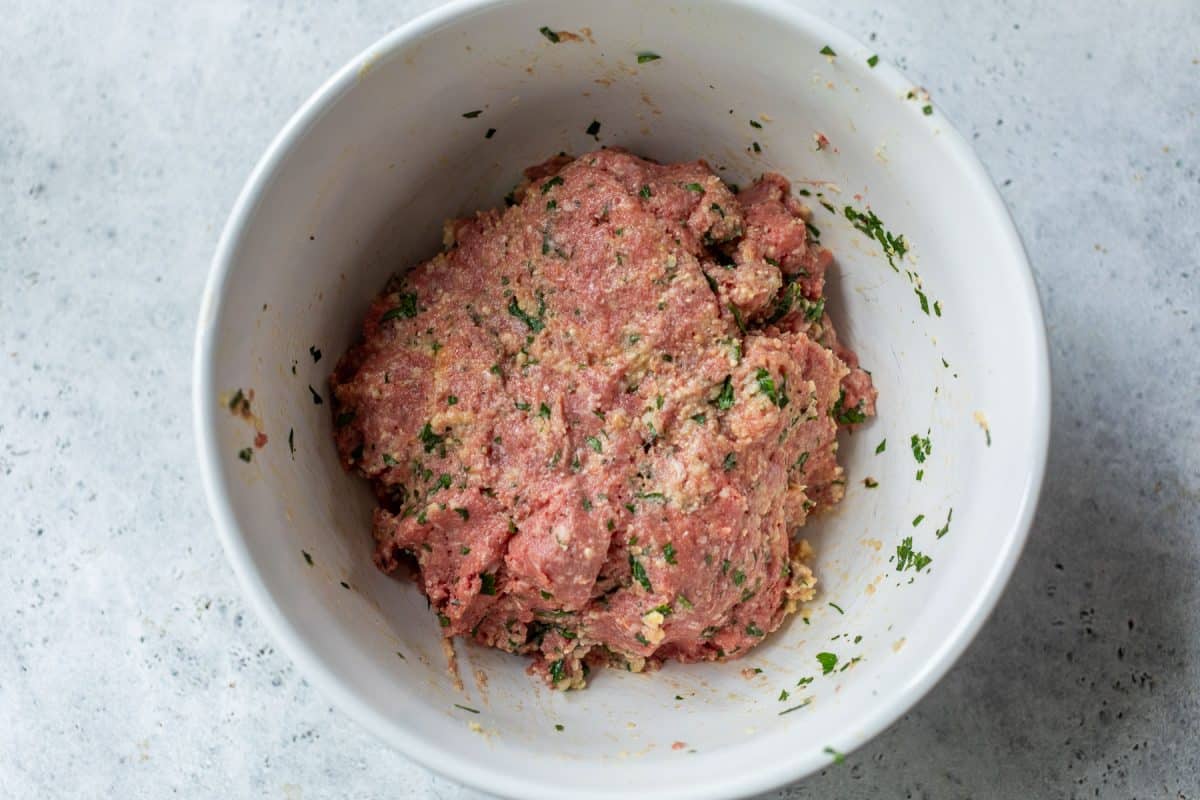 Ground meat mixed with herbs and spices in a white bowl on a gray surface.