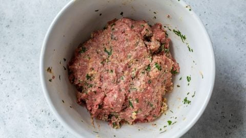 Ground meat mixed with herbs and spices in a white bowl on a gray surface.