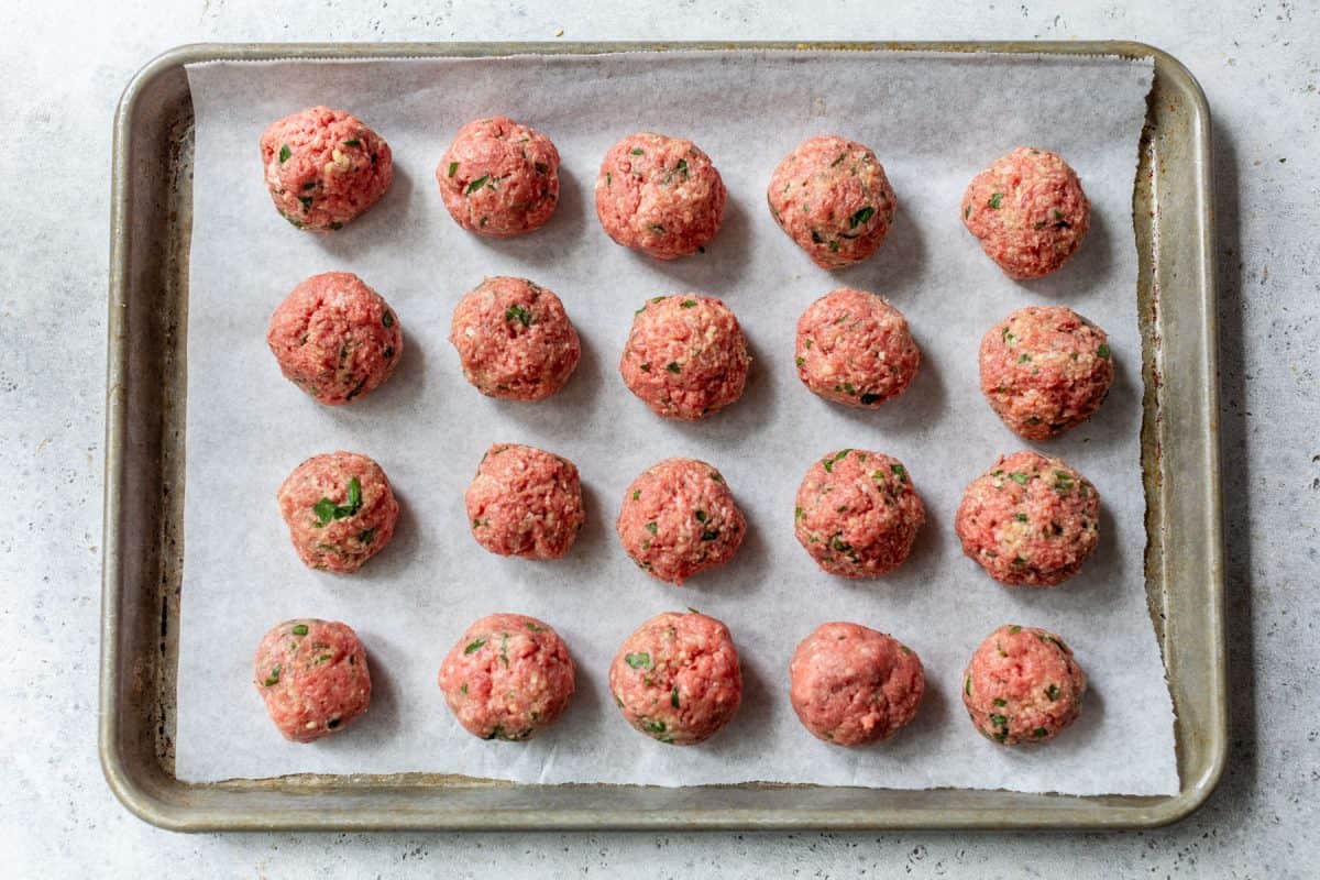 Uncooked ground beef meatballs arranged in rows on a parchment-lined baking sheet.