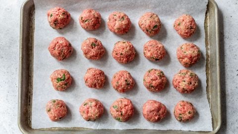 Uncooked ground beef meatballs arranged in rows on a parchment-lined baking sheet.