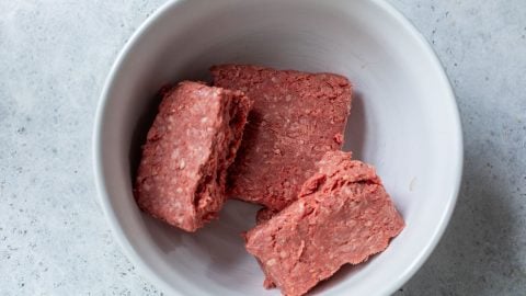 Three rectangular chunks of raw ground beef in a white bowl on a light gray countertop.