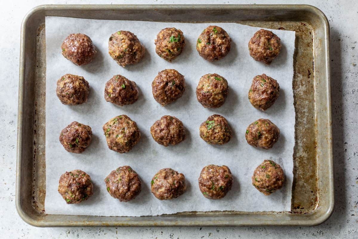 Twenty baked ground beef meatballs arranged in rows on parchment paper on a baking sheet.