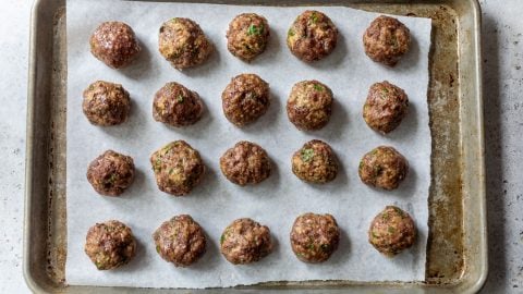 Twenty baked ground beef meatballs arranged in rows on parchment paper on a baking sheet.