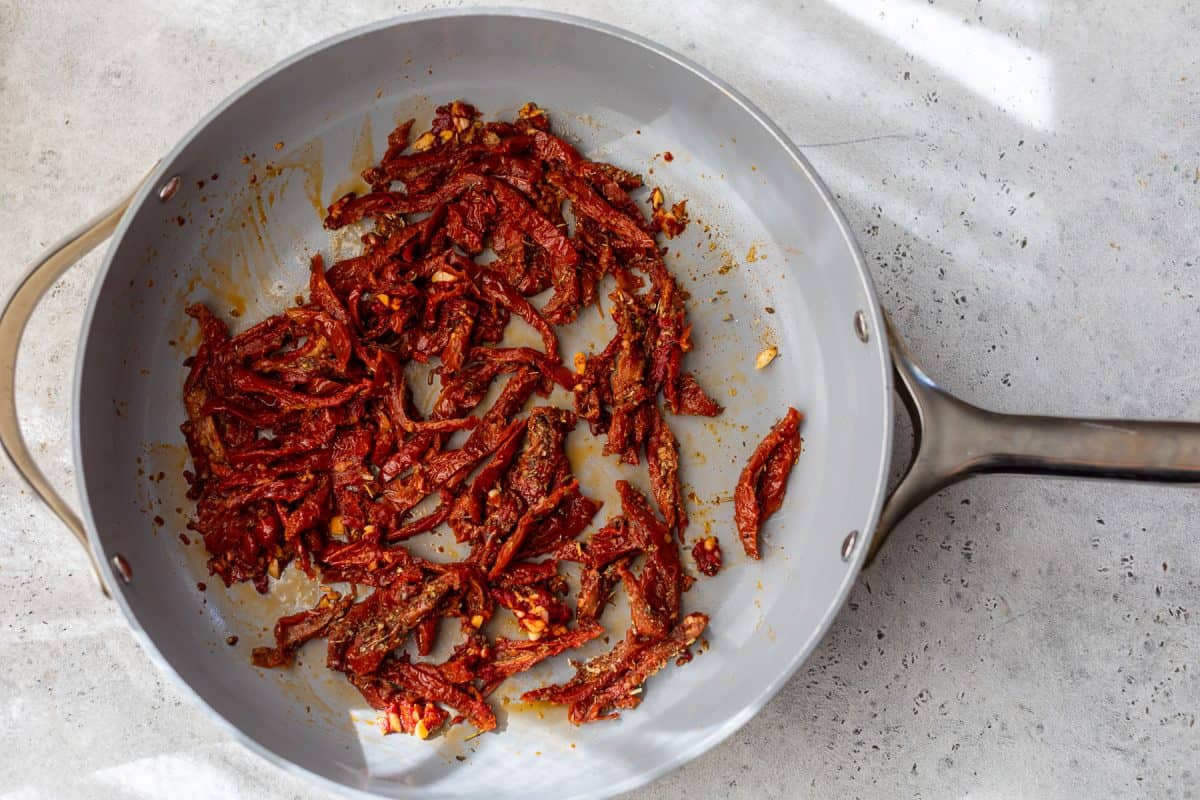 Sliced sun-dried tomatoes sautéing in a gray frying pan on a light-colored countertop.