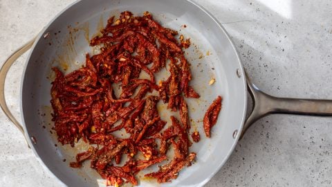 Sliced sun-dried tomatoes sautéing in a gray frying pan on a light-colored countertop.