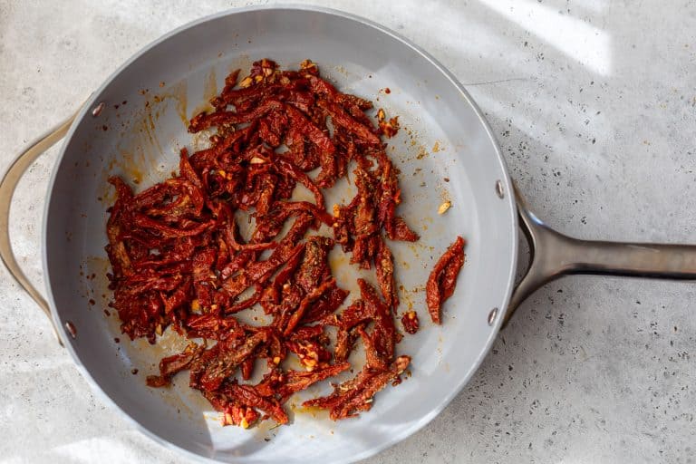 Sliced sun-dried tomatoes sautéing in a gray frying pan on a light-colored countertop.