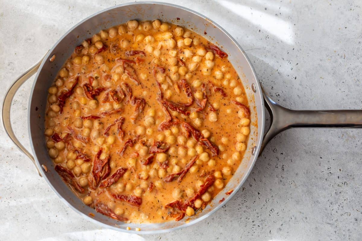 Creamy chickpea stew with sun-dried tomatoes in a skillet on a light countertop, seen from above.