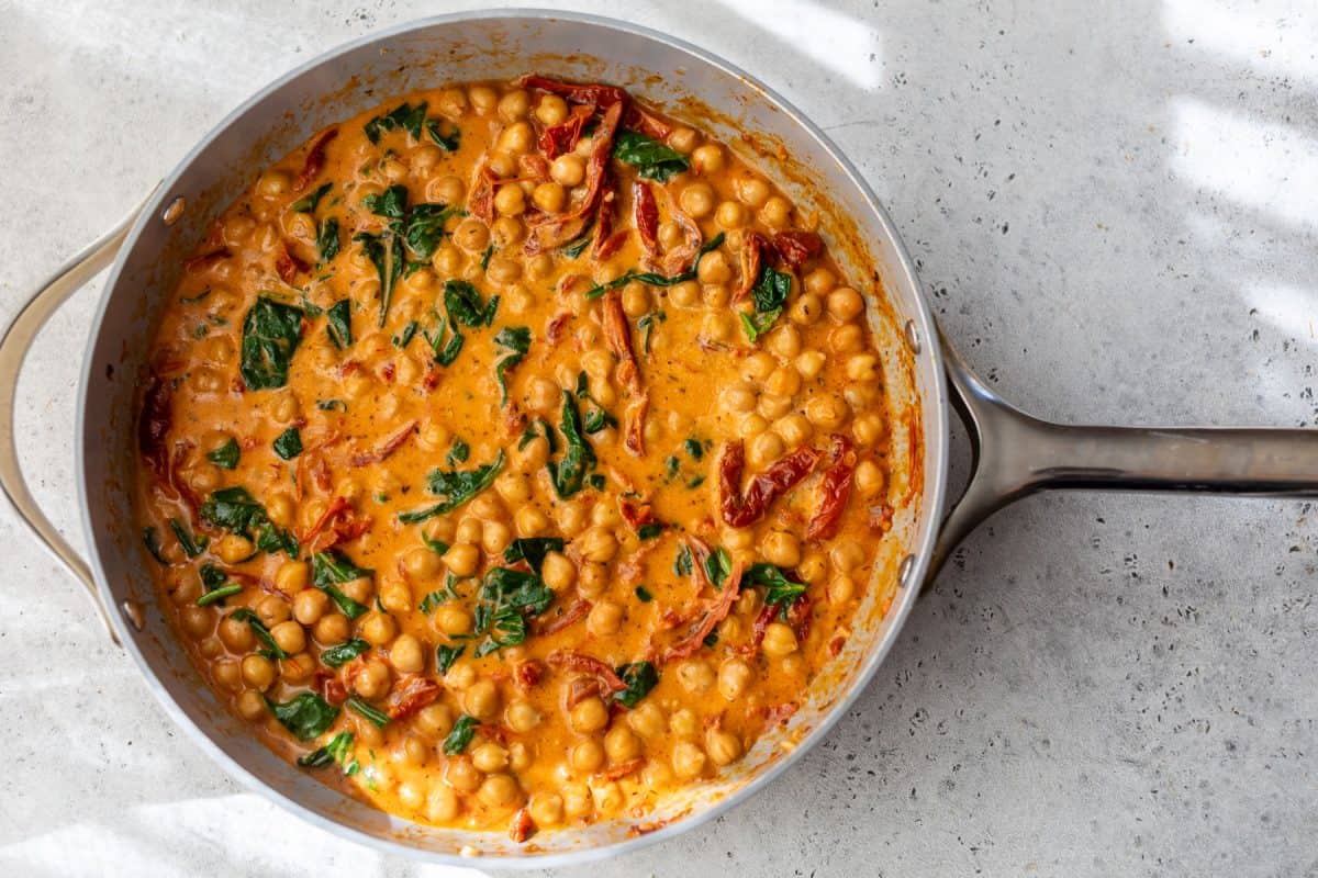 A pan of creamy chickpea stew with spinach and sun-dried tomatoes in a rich orange sauce on a light countertop.