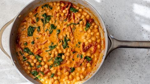 A pan of creamy chickpea stew with spinach and sun-dried tomatoes in a rich orange sauce on a light countertop.