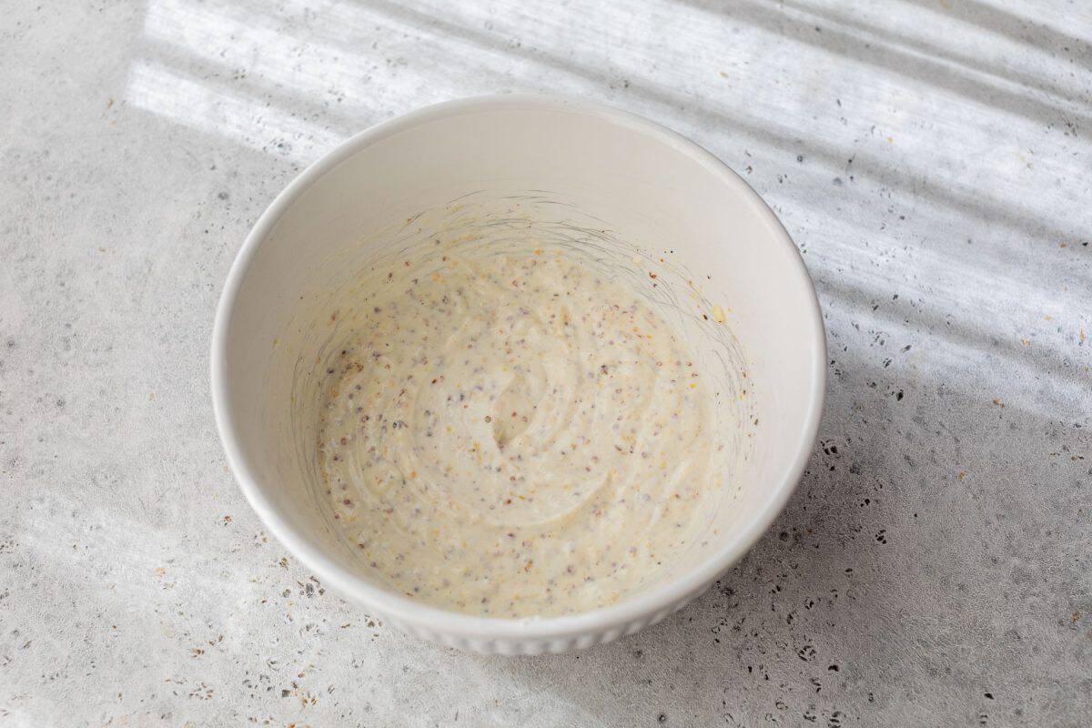 A white bowl of creamy potato salad dressing with mustard seeds sits on a light textured surface in natural light.