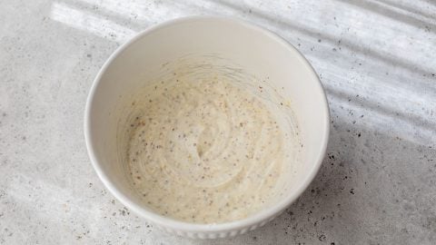 A white bowl of creamy potato salad dressing with mustard seeds sits on a light textured surface in natural light.
