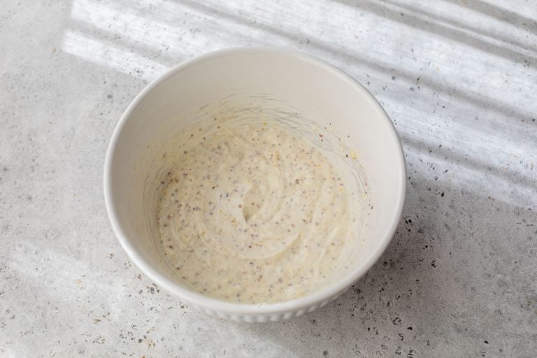 A white bowl of creamy potato salad dressing with mustard seeds sits on a light textured surface in natural light.