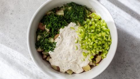 A bowl with chopped potatoes, celery, fresh herbs, and creamy dressing on a light surface.