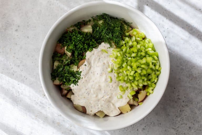 A bowl with chopped potatoes, celery, fresh herbs, and creamy dressing on a light surface.