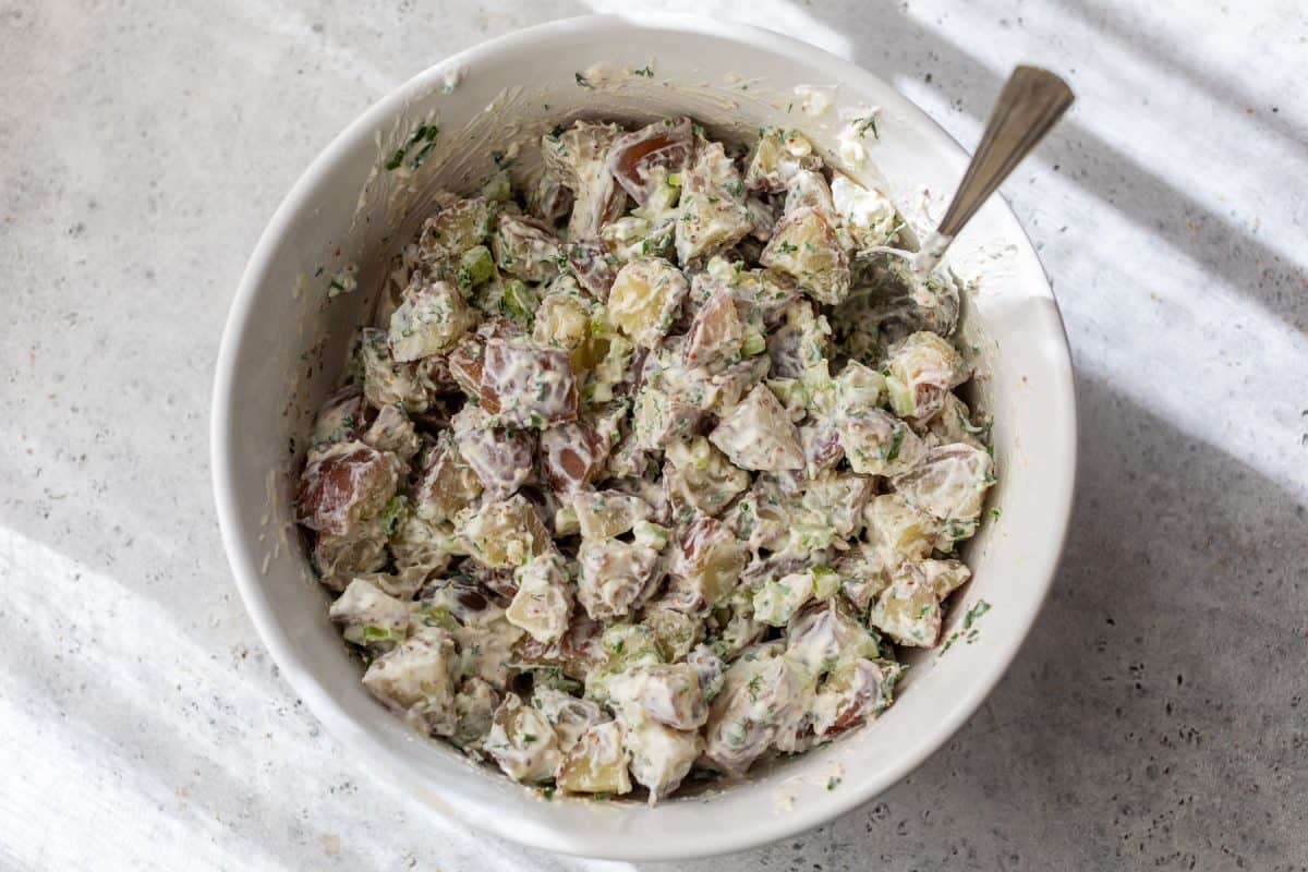 A bowl of creamy red potato salad with herbs and chopped vegetables, with a spoon inside the bowl.
