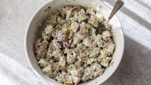 A bowl of creamy red potato salad with herbs and chopped vegetables, with a spoon inside the bowl.