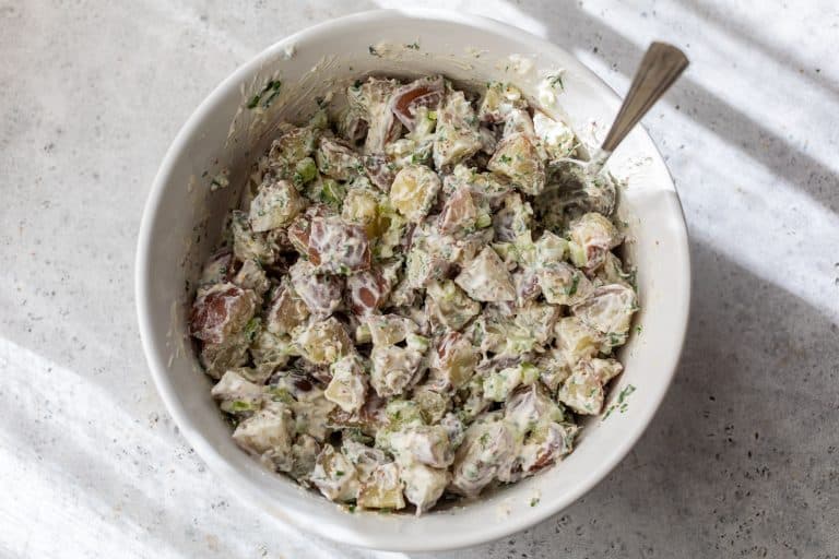 A bowl of creamy red potato salad with herbs and chopped vegetables, with a spoon inside the bowl.