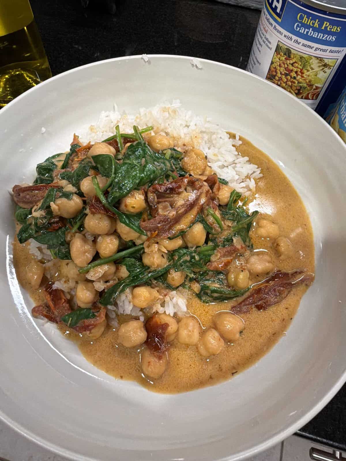 White bowl with rice topped with chickpeas, spinach, sun-dried tomatoes, and creamy sauce; canned chickpeas in background.
