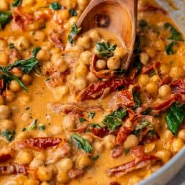 Creamy chickpea stew with sun-dried tomatoes and spinach being stirred with a wooden spoon in a pan.