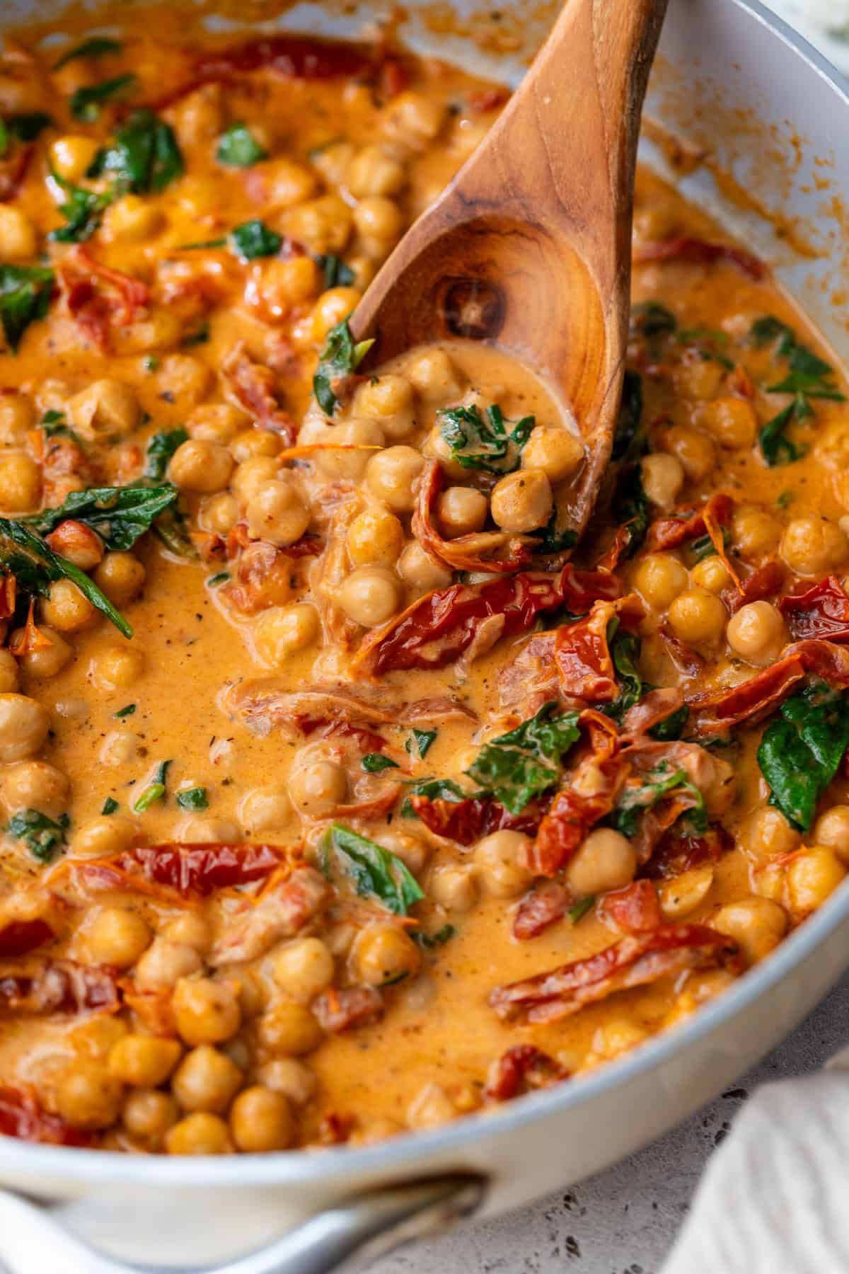 Creamy chickpea stew with sun-dried tomatoes and spinach being stirred with a wooden spoon in a pan.