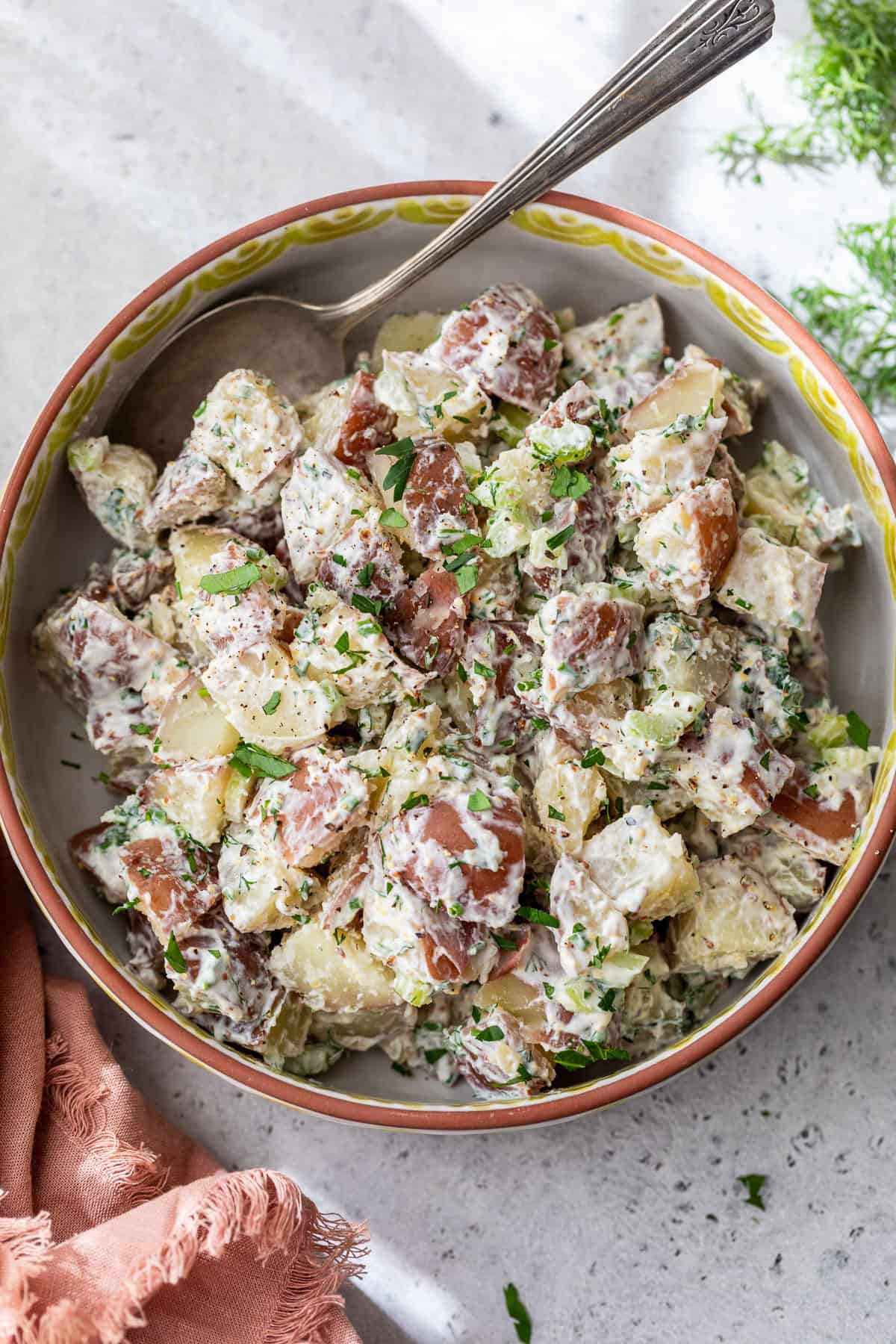 Bowl of creamy red potato salad with chopped herbs, red-skinned potatoes, and a spoon on a light surface.
