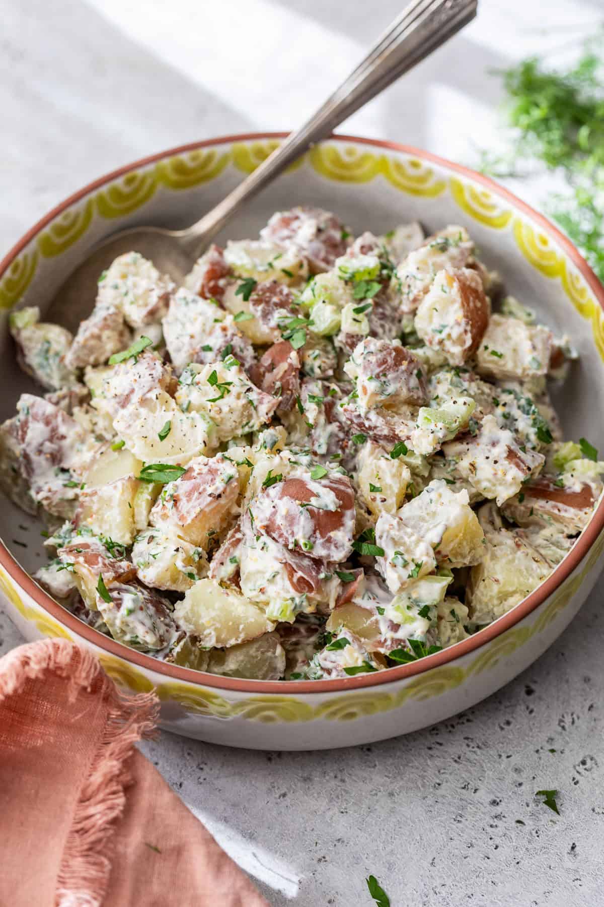 A bowl of creamy red potato salad with herbs and a spoon, placed on a light-colored surface.
