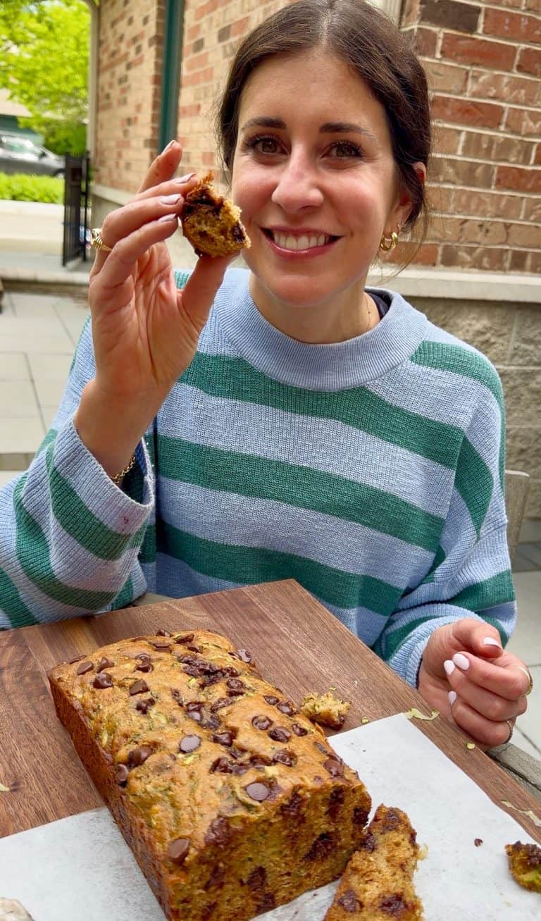 Erin Clarke smiles while holding a piece of chocolate chip zucchini bread, with a loaf on the table in front of her.