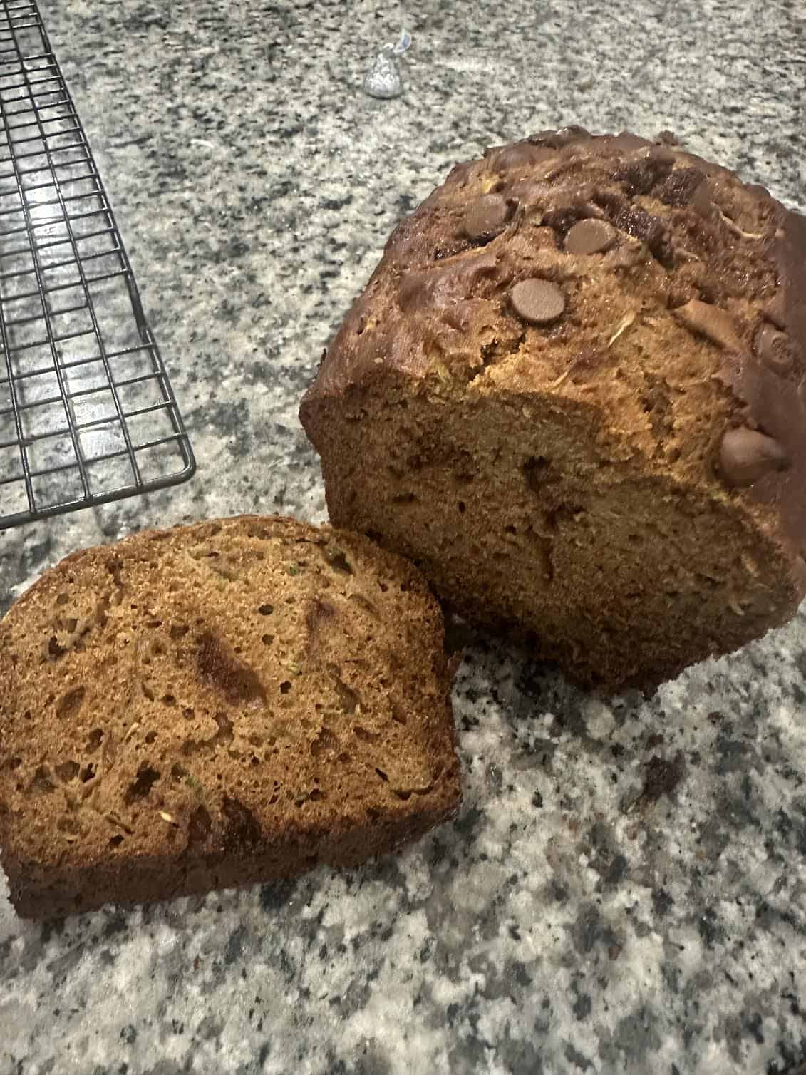A loaf of bread with one slice cut, sitting on a granite countertop next to a cooling rack.