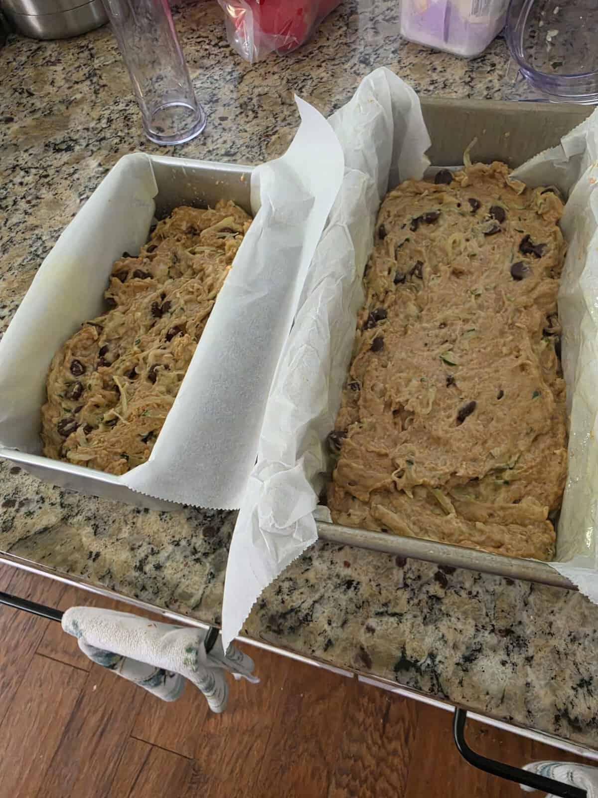 Two loaf pans with unbaked batter, some with chocolate chips, on a granite kitchen counter lined with parchment paper.