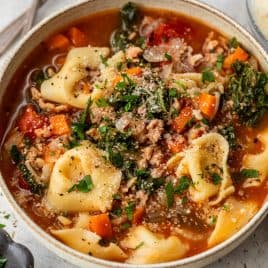 Bowl of crockpot tortellini soup with vegetables, ground meat, greens, and grated cheese, garnished with fresh herbs.