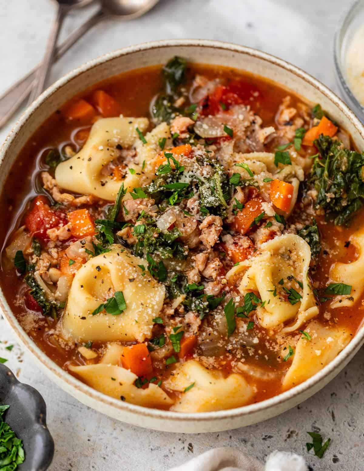 Bowl of crockpot tortellini soup with vegetables, ground meat, greens, and grated cheese, garnished with fresh herbs.