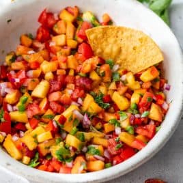 A bowl of peach salsa with diced tomatoes, onions, cilantro, and a tortilla chip dipped inside.
