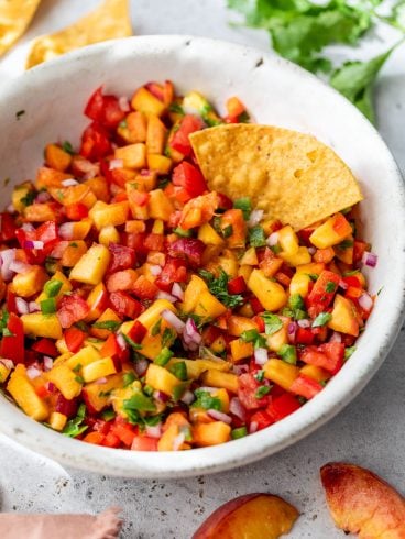 A bowl of peach salsa with diced tomatoes, onions, cilantro, and a tortilla chip dipped inside.