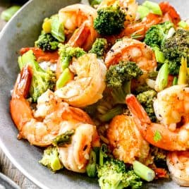 Shrimp stir-fry with broccoli and green onions served in a gray bowl on a wooden table.
